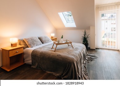 Small attic bedroom in a family house with morning breakfast service on the bed. The bed also has a fluffy blanket and two fluffy pillows, on the sides there are bedside tables with lit lamps. - Powered by Shutterstock