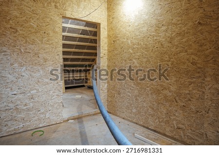 Small attic room lined with OSB shows doorway to sloped roof cavity with exposed rafters, insulation wrap labeled TOP, gray hose on subfloor, and green cable loop.