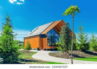A small barnhouse house on a plot with a path, trees and bushes under a blue sky