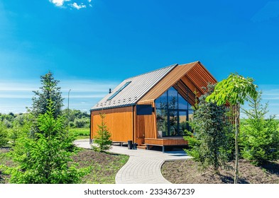 A small barnhouse house on a plot with a path, trees and bushes under a blue sky