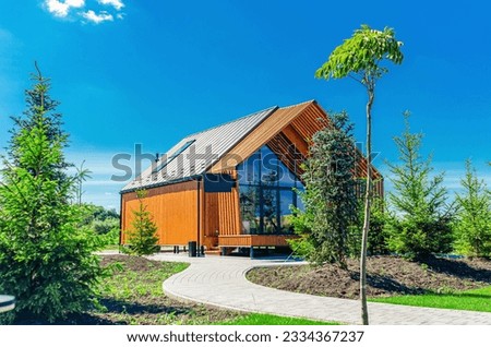 A small barnhouse house on a plot with a path, trees and bushes under a blue sky