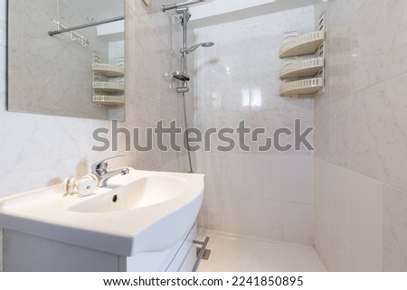 Small bathroom with a white porcelain sink on a wooden cabinet and a frameless square mirror, a plastic shelf in one corner and a walk-in shower without a screen