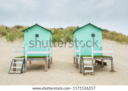 small beach houses with steps and a gate with in the background dunes with dune grass under a cloudy sky