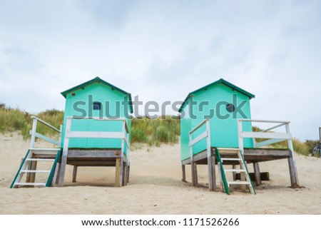 small beach houses with steps and a gate with in the background dunes with dune grass under a cloudy sky