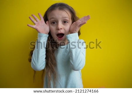 Small beautiful girl puts arms up and screams from joy. Child with long hair dressed in pale blue t-shirt on yellow background. Emotional kid.