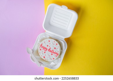 A small bento birthday cake in a box with a wooden spoon. Yellow and pink background