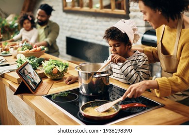 Small Black Boy Cooking With Is Mother In The Kitchen. Father And Daughter Are In The Bakground. 