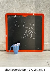 Small black drawing board with white chalk writing displaying basic math and alphabet lessons, accompanied by a blue chalk holder, hanging on a textured white wall.