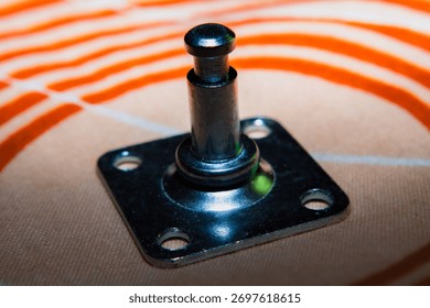 A small black object with a silver screw on top sits on a tan surface. The object is square in shape and has a black and silver color scheme.