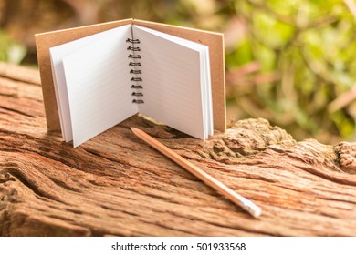 Small blank note book open and pencil on old wooden texture with green nature background. Closeup, Select focus.