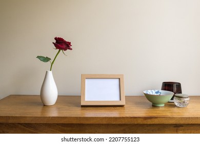 Small Blank Picture Frame On Oak Side Table With Single Red Rose And Bowls Against Beige Wall