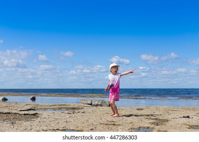 small blond girl wearing pink clothing and cap standing on rocky sea beach with shallow water and pointing away with finger  - Shutterstock ID 447286303