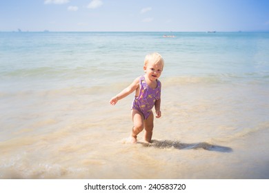 small blonde baby girl in purple swim suit play with waves on seashore