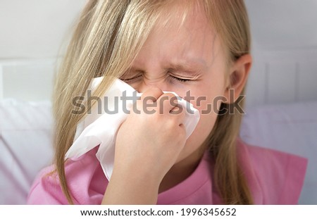 A small blonde girl with blond hair, white skin, European appearance blows her nose in a white kerchief. The child has closed his eyes and sneezes. Influenza, infection, allergy, virus, cold, runny