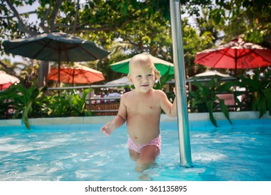 Small Blonde Girl Smiles And Holds Pole In Shallow Water Of Hotel Swimming Pool Against Colourful Shade Parasols