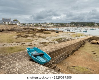 A small blue boat rests on a stone pier at low tide, surrounded by a coastal landscape. A view of the marina and village of Ploumanach - Powered by Shutterstock