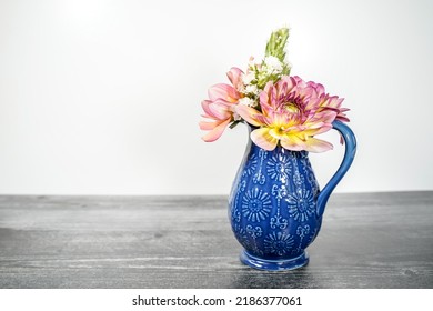 Small Blue Ceramic Vase Filled With Two Pink Dahlia Flowers And A Variety Of Freshly Cut Flowers From A Garden. White Background And Weather Black Wood Base.