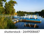 Small blue ferry docked on a calm river with green trees and grass at sunset. Peaceful summer landscape, reflection in water, rural nature, and transportation scene in the countryside.