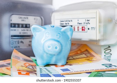 Small blue piggy bank and Euro cash near an electricity meter and gas meter. Utility bills, consumption of electricity and gas for heating home, energy costs, symbolic image.