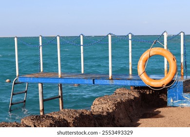 small blue pontoon with iron pylons for barriers on the sides, a ladder to descend into the sea, lifebuoy, horizon with blue sky and blue sea - Powered by Shutterstock