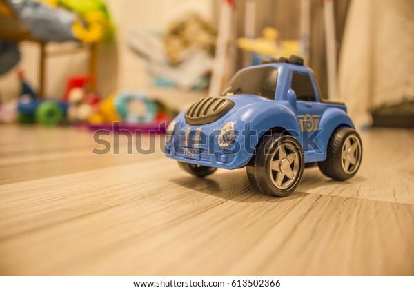 Small blue toy car on the wooden\
floor. Stands in front of other toys and looks to the left.\
