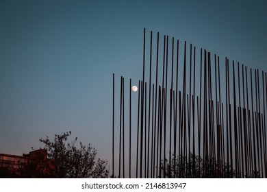 Small Blurry Moon Almost Full Circle In Blue Sky Between Construction Site Bars And Tree Crown