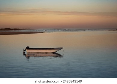 A small boat floats peacefully on calm water at sunset, with a vibrant orange sky reflecting off the water's surface and a distant shoreline silhouetted against the horizon. - Powered by Shutterstock