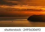 A small boat slithers through the calm waters of Possession Sound and Puget Sound. The sky is painted in warm hues of orange and red, with mountains of the Olympic Peninsula visible in the distance.