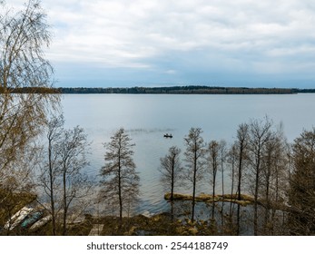 A small boat with two people floats on a calm lake, surrounded by bare trees. An overcast sky casts muted light, with a forest visible in the distance. - Powered by Shutterstock