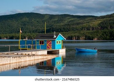 A small boathouse at the end of a wooden pier. The bright teal blue building has a red door and yellow trim. There's a small rowboat anchored to the wharf on a pond with a hill in the background.