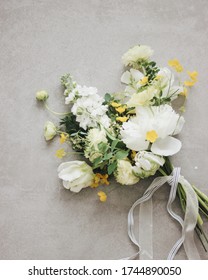 A Small Bouquet Of White And Yellow Spring Flowers (ranunculus/buttercup, Tulip, Peony, Matthiola, Spirea) With Beautiful Ribbons Laying On The Gray Concrete Textured Background. High Angle View. Spac