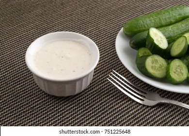 A Small Bowl Of Ranch Dressing With Sliced And Whole Bite Size Cucumbers On A White Plate Plus A Fork In The Foreground On A Tablecloth.