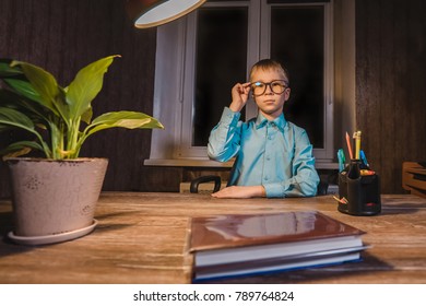 Small boy in glasses sitting at table with lamp, books, plant and pens.  
