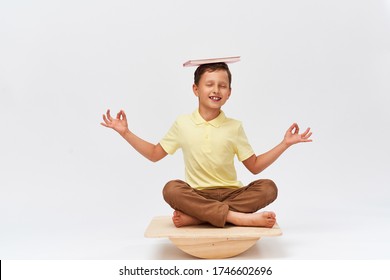 Small Boy Holds Book On His Head While Balancing On Special Simulator For Training Muscles. Child Is Engaged On Balance Beam, An Intermediate Internal Balance. Training Development Cerebellum.