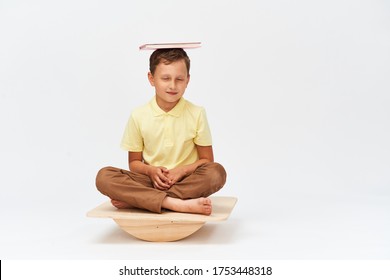 Small Boy Holds Book On His Head While Balancing On Special Simulator For Training Muscles. Child Is Engaged On Balance Beam, An Intermediate Internal Balance. Training Development Cerebellum.