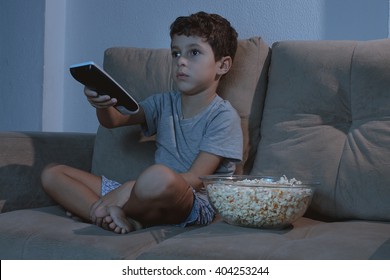 Small Boy On The Couch Watching TV And Eating Popcorn At Night In The Living Room