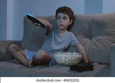 Small Boy On The Couch Watching TV And Eating Popcorn At Night In The Living Room