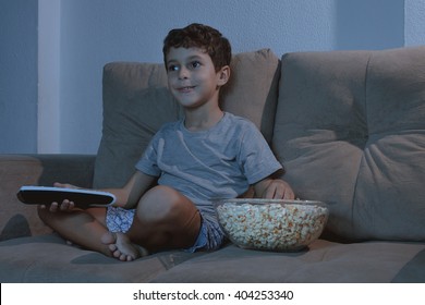 Small Boy On The Couch Watching TV And Eating Popcorn At Night In The Living Room