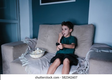 Small Boy On The Couch Watching TV And Eating Popcorn At Night In The Living Room