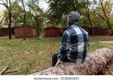 A small boy in a plaid shirt sits on a felled tree against an old iron garages - Powered by Shutterstock