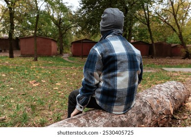 A small boy in a plaid shirt sits on a felled tree against an old iron garages - Powered by Shutterstock