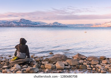 Small Boy Sits On Rock By Okanagan Lake With Sunset View Of Snow Covered Mountains