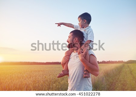 Small boy is sitting on his father??s shoulders and pointing at the sun in the field during beautiful sunset 