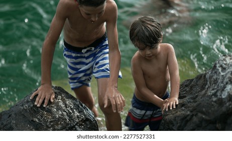 Small boy standing by rocks shore at lake water. Brave wet child stands outdoors holding on large stone