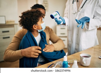 Small boy wearing face mask and sitting in father's lap while doctor is measuring his temperature during home visit. 