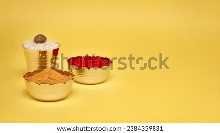Small brass bowl with Sindoor or kumkum, Haldi, and a brass glass with rice and betel nut (supari). Hindu pooja objects over a yellow background.