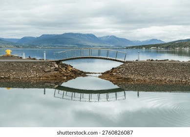 A small bridge forming a path next to the port of Akureyri. The fjord with water like a mirror and the mountains on the background.