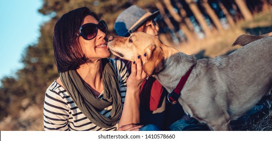 Small Brown Dog Licking Woman Face While Resting On Grass By The Pine Forest