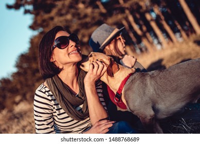 Small Brown Dog Licking Woman Face While Resting On Grass By The Pine Forest