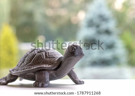 a small brown iron turtle on a white surface by the window in front of the garden on a sunny day there is a shape of fir trees, idea of slow living, enjoying countryside lifestyle, artificial animal